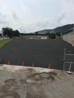 A newly paved, dark asphalt surface sits in front of a long industrial building, lined with four orange traffic cones.