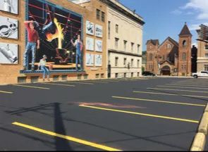 A parking lot in front of a brick building featuring a large mural of people working with molten metal and various tools.