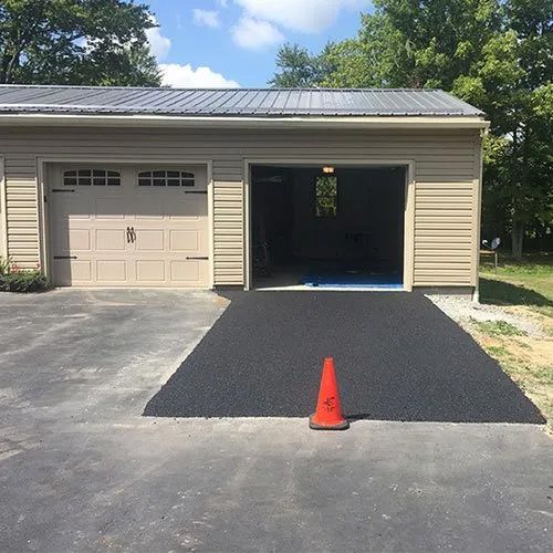 A detached garage with one closed and one open door, with fresh dark asphalt in front of the open bay and a traffic cone.