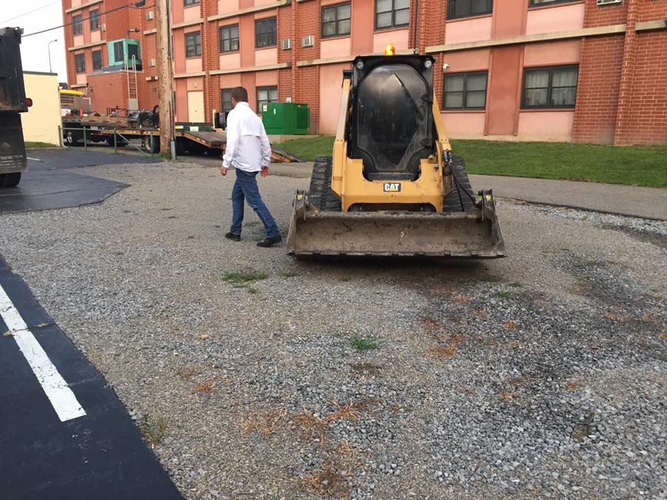 A person in a white jacket and jeans walks past a yellow Caterpillar skid steer on a gravel lot near a brick building.