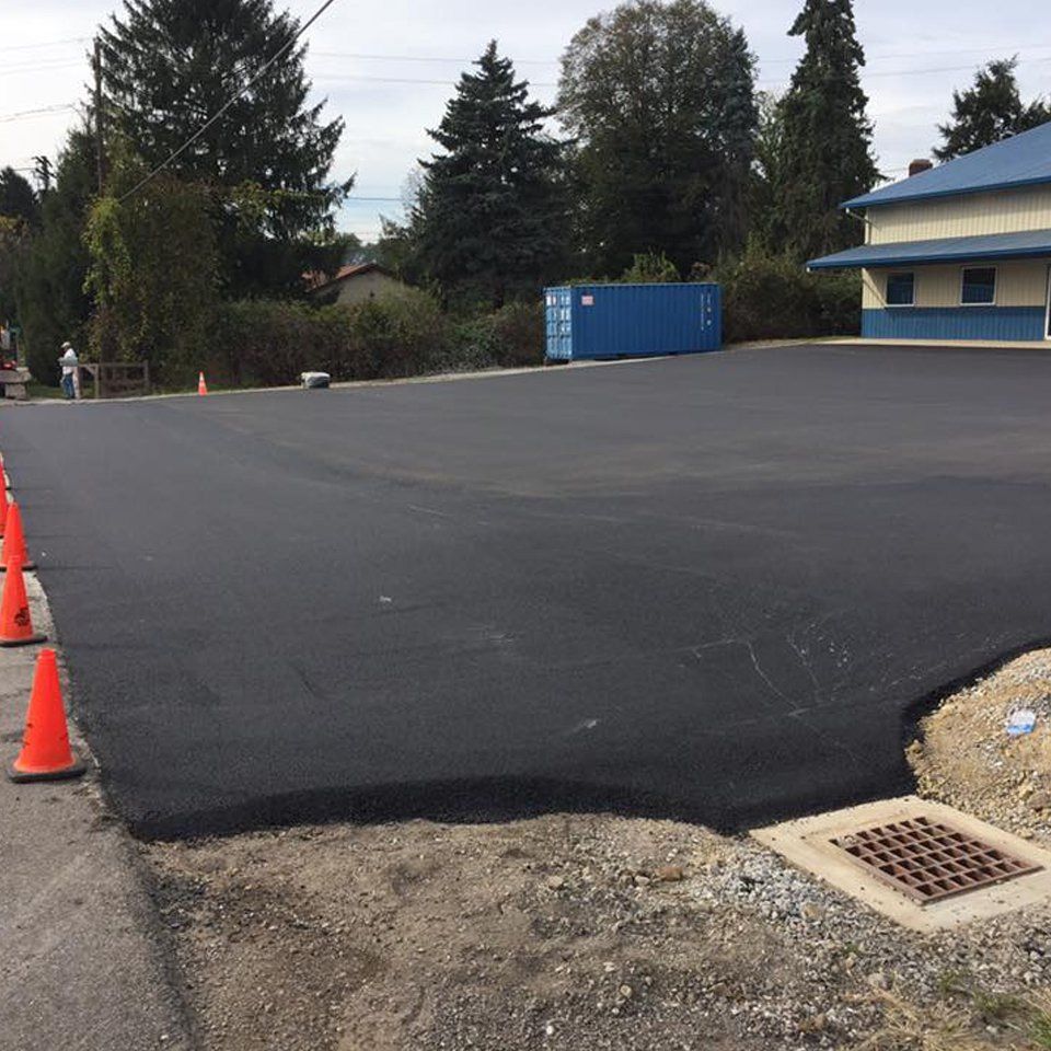 A newly paved asphalt parking lot sits next to a blue building and a blue storage container, with orange safety cones.