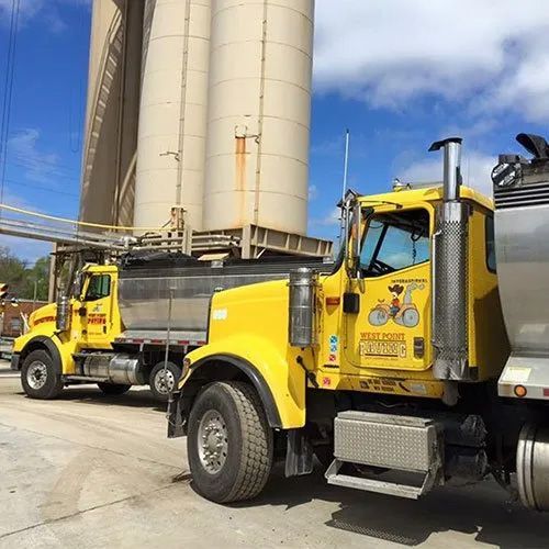Two yellow dump trucks are parked side by side under large industrial silos against a blue sky.