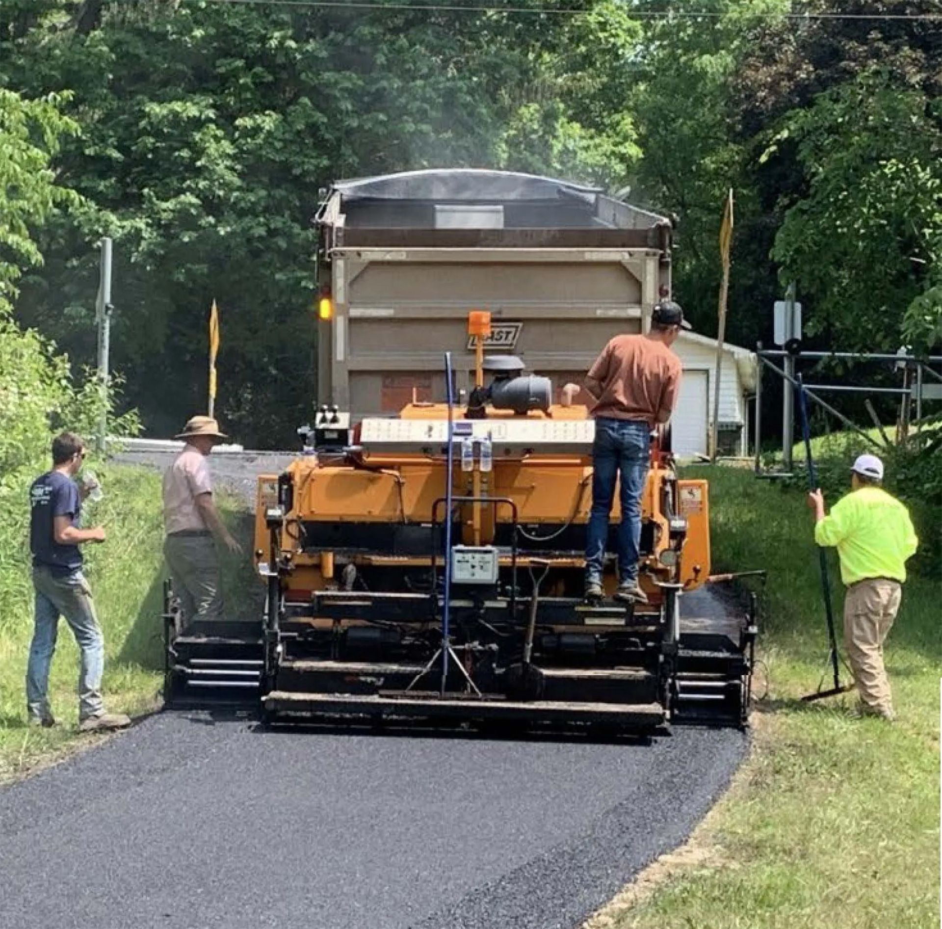 A road paving crew operates a large yellow paver machine on a fresh asphalt surface surrounded by trees.