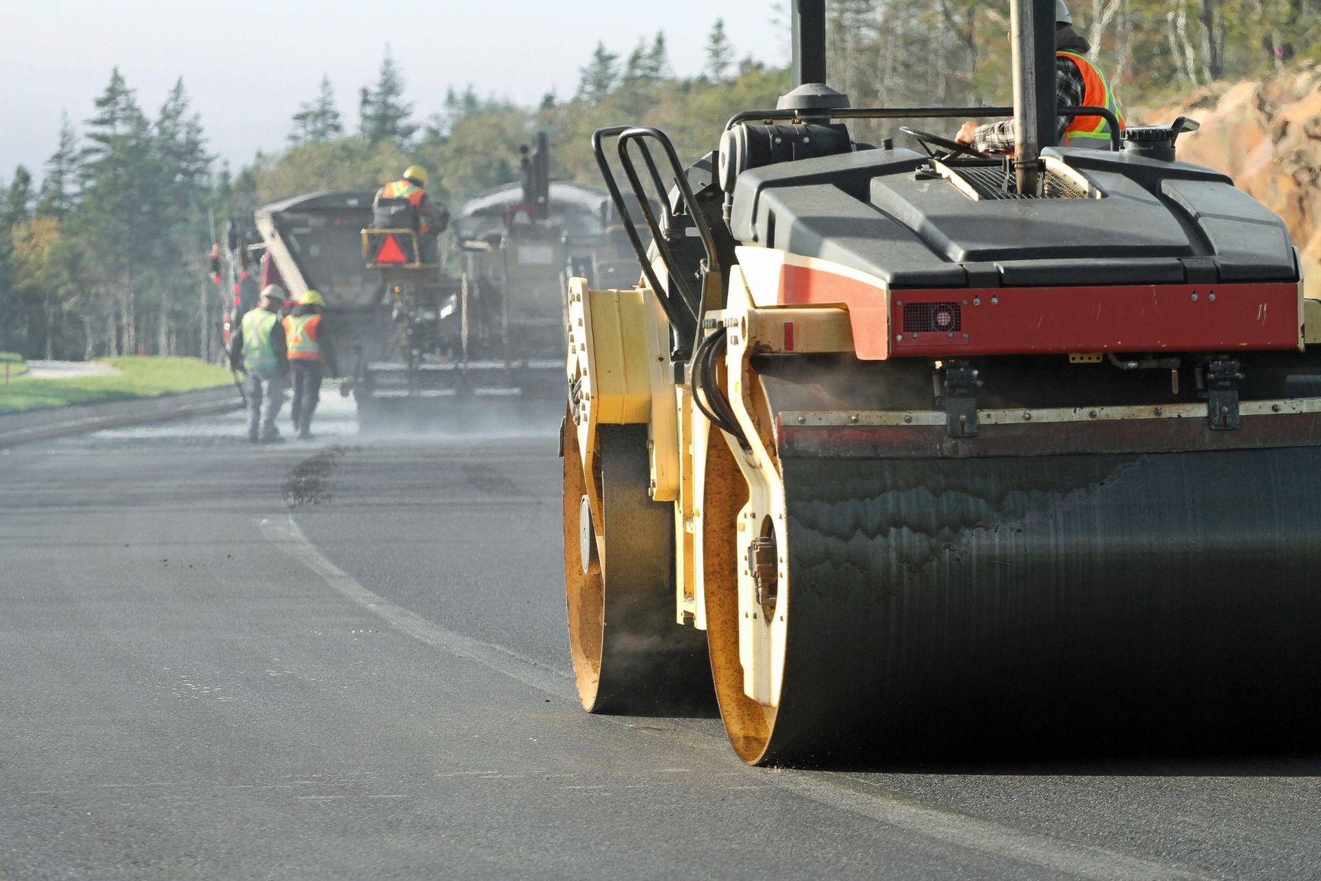 A yellow road roller works on a freshly paved asphalt road with construction workers and machinery in the background.