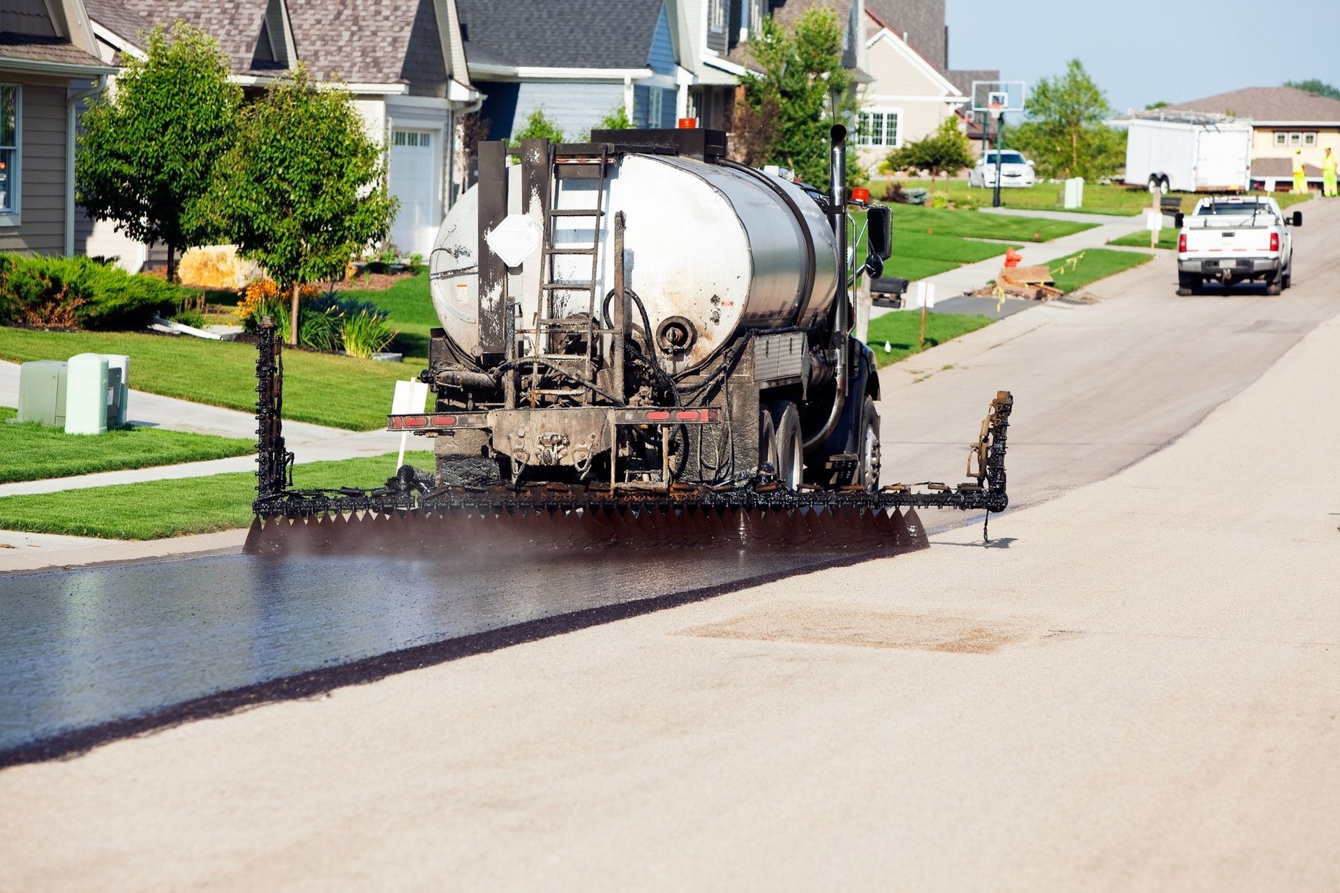 A truck applies a dark sealcoat to a residential street, leaving a freshly coated surface behind it.