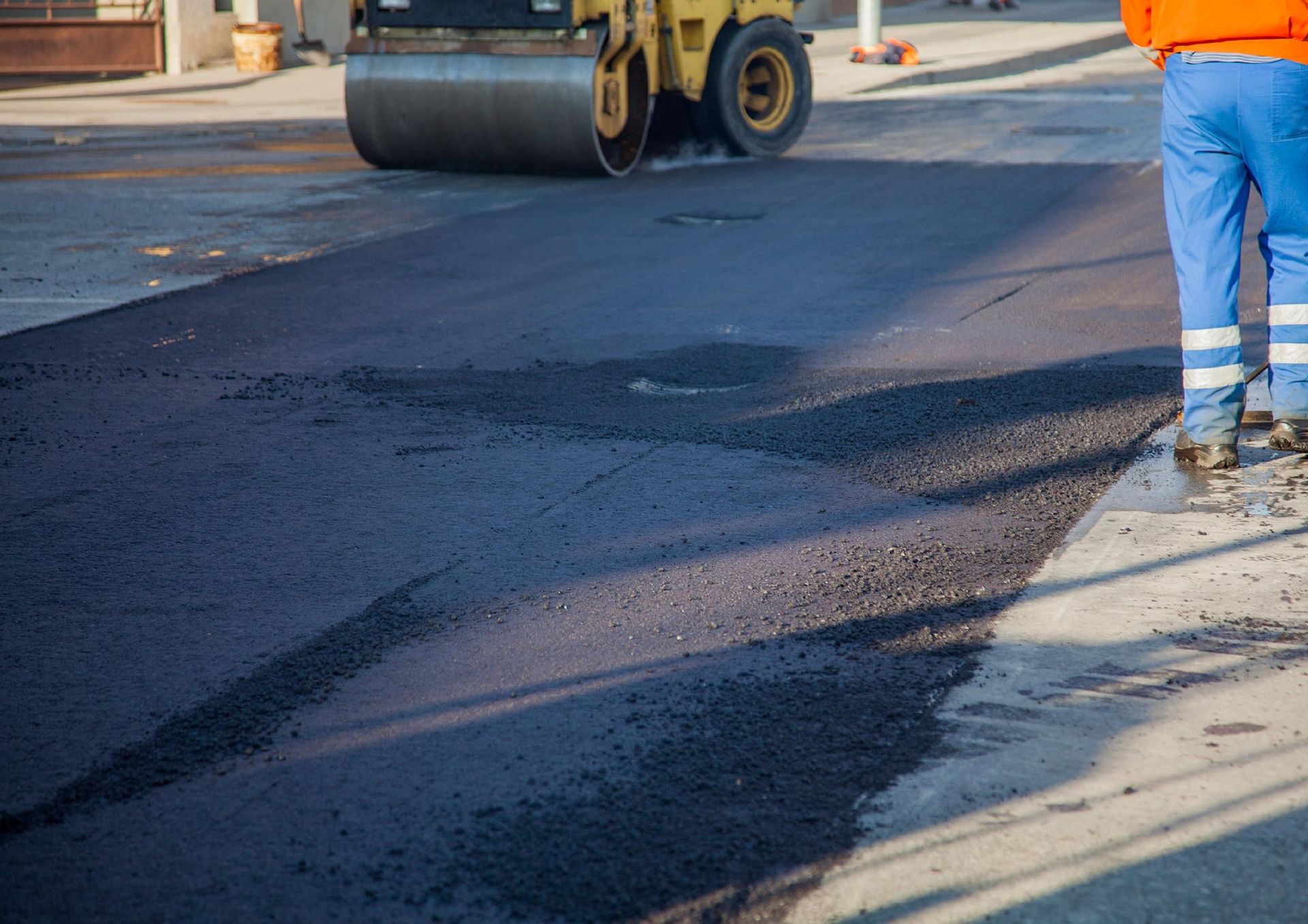 A road roller compacts freshly laid black asphalt on a street as a worker in blue pants stands nearby.