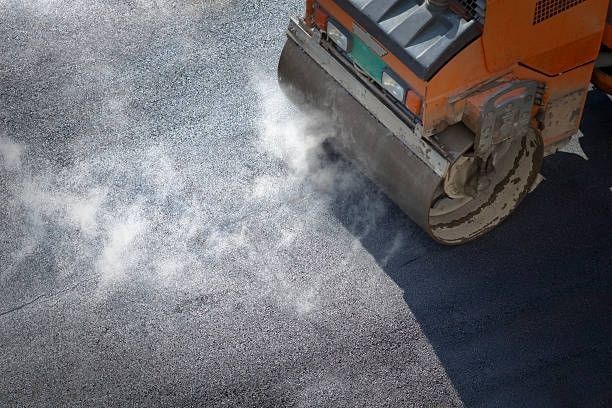 An orange road roller compacts freshly laid, steaming hot asphalt on a construction site.