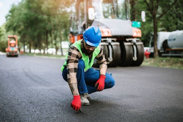 A worker in a high-visibility vest and blue hard hat crouches on newly paved asphalt, inspecting the surface.