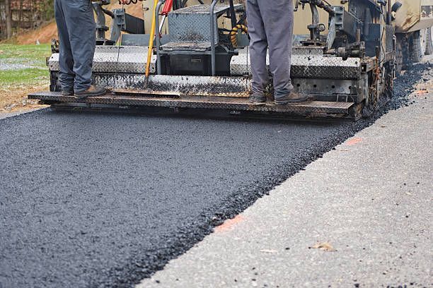 Two workers stand on the back of a paving machine as it lays fresh black asphalt onto a road.
