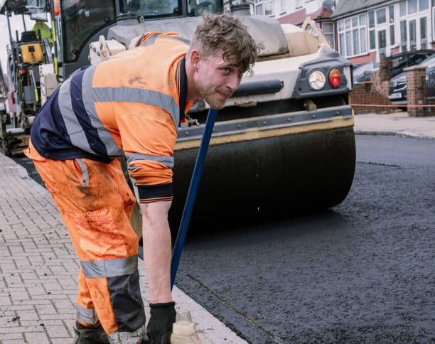 A worker in high-visibility orange clothing uses a tool to smooth asphalt near a large road roller on a residential street.