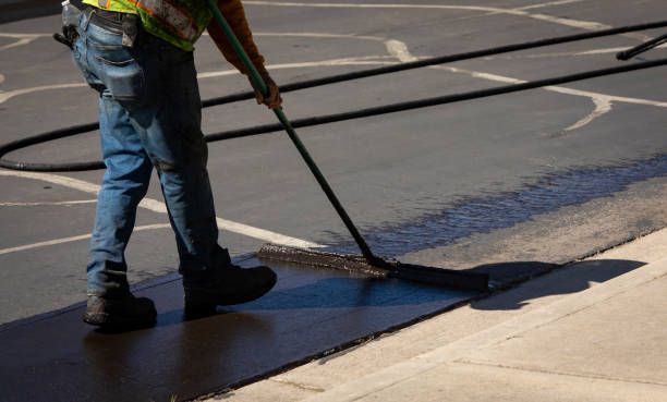A worker in a high-visibility vest uses a long-handled tool to apply dark sealant to a cracked asphalt parking lot.