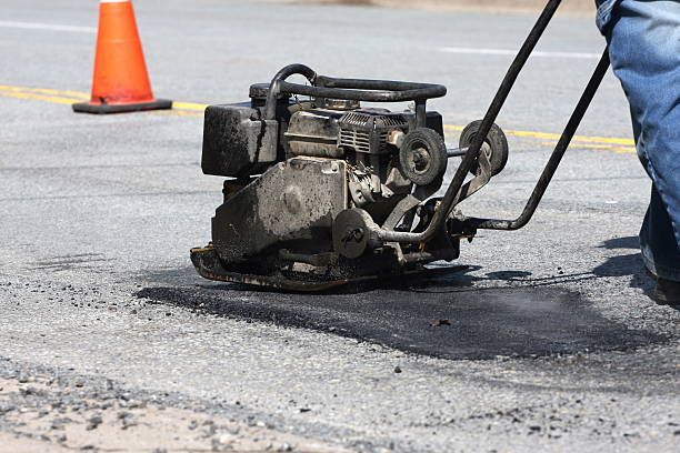 A worker uses a vibrating plate compactor to smooth fresh asphalt patch on a road surface near an orange traffic cone.