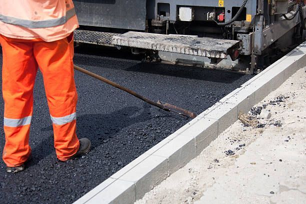 A worker in high-visibility orange clothing uses a rake to spread fresh black asphalt next to a concrete curb.