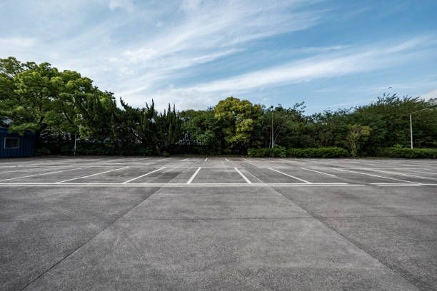 An empty asphalt parking lot with painted white spaces against a backdrop of trees and a blue, cloudy sky.