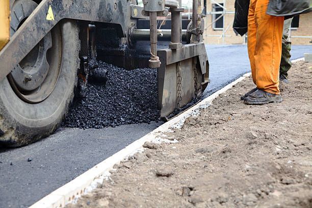 A paving machine lays fresh, dark asphalt along a concrete curb next to a worker in orange trousers.