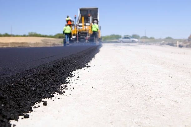 Workers operate a paving machine to lay fresh black asphalt onto a light-colored road base under a clear blue sky.