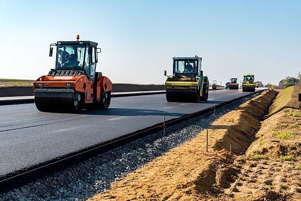 Orange and yellow construction rollers compact a new asphalt road under a clear blue sky.