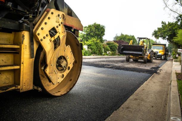 A yellow road roller works on a suburban street being repaved, with a front-loader and dump truck in the background.