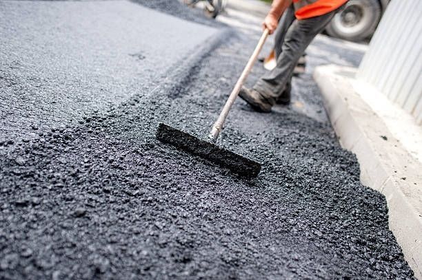 A worker in high-visibility gear uses a rake to spread freshly laid, dark asphalt on a road construction site.