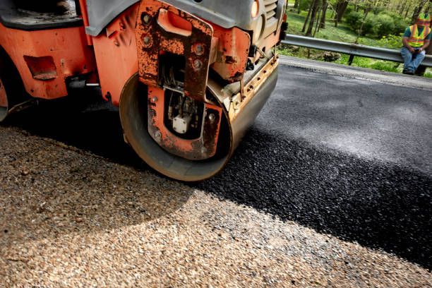 An orange road roller compacts freshly laid black asphalt on a road, with a construction worker visible in the background.