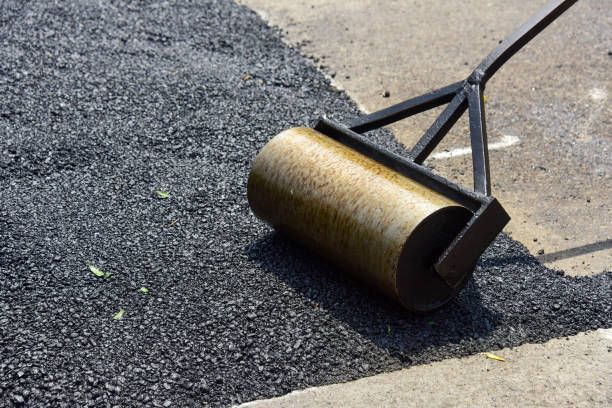 A handheld metal lawn roller flattening fresh, dark asphalt on a paved surface.