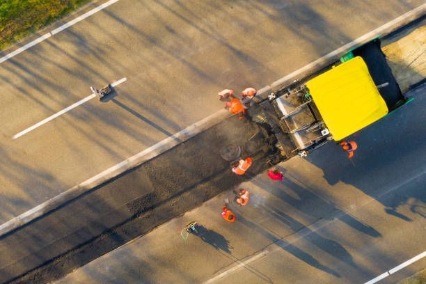 An aerial view of a yellow road paver laying fresh asphalt on a highway, with workers in orange vests working alongside it.