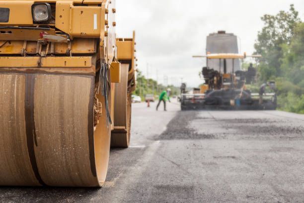 A yellow road roller occupies the foreground, with construction equipment and a worker visible on a paved road beyond.