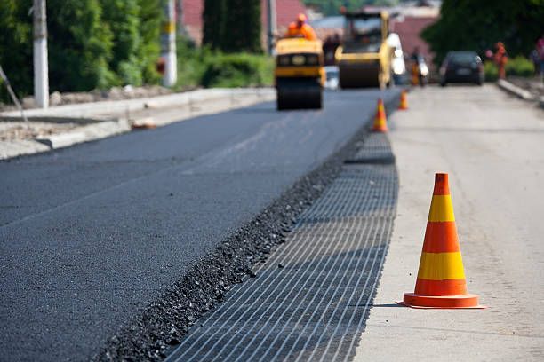 Construction workers use heavy road rollers to pave a new black asphalt road marked by bright orange and yellow traffic cones.