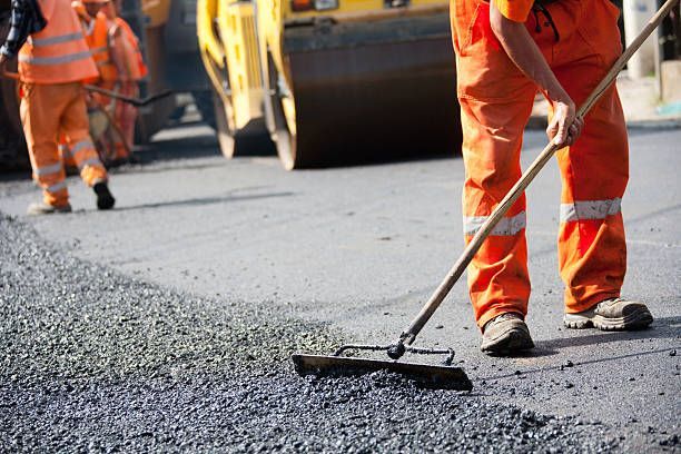 Construction workers in high-visibility orange uniforms use tools to pave a road with asphalt as a steamroller approaches.