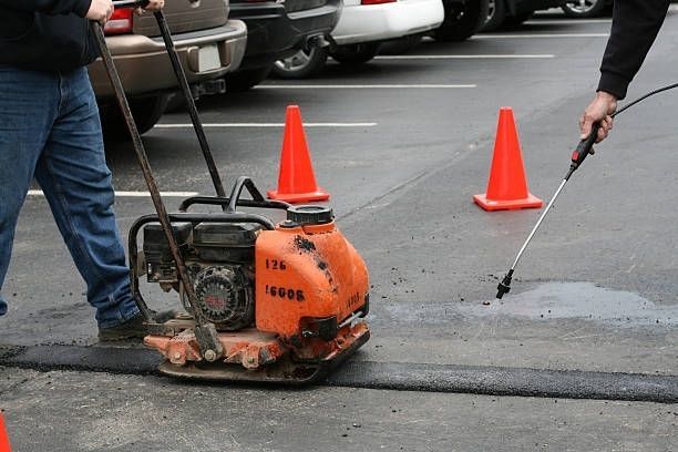 Workers use a vibratory plate compactor and a spray wand to patch and seal asphalt pavement in a parking lot.