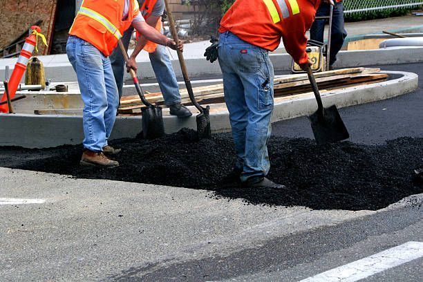 Construction workers in orange safety vests use shovels to spread hot asphalt on a road surface.