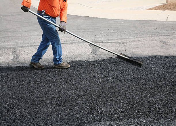 A construction worker in an orange shirt uses a long-handled rake to smooth fresh asphalt on a road.