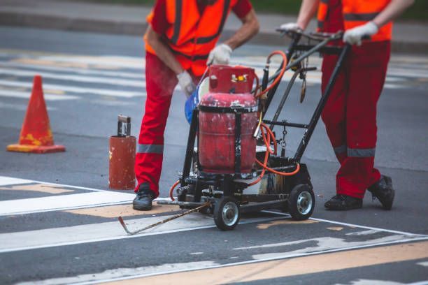 Two workers in bright orange safety vests and red pants use a road marking machine to paint a crosswalk.