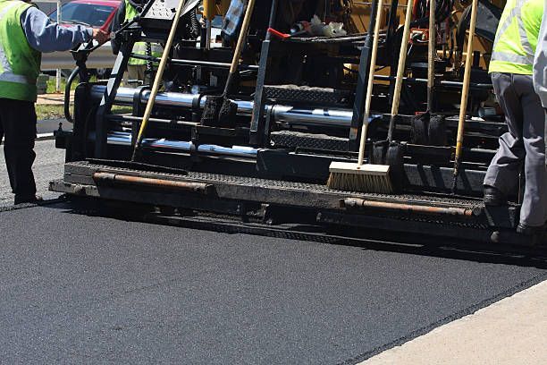 Two workers in high-visibility vests operate a paving machine to lay fresh asphalt on a road surface.