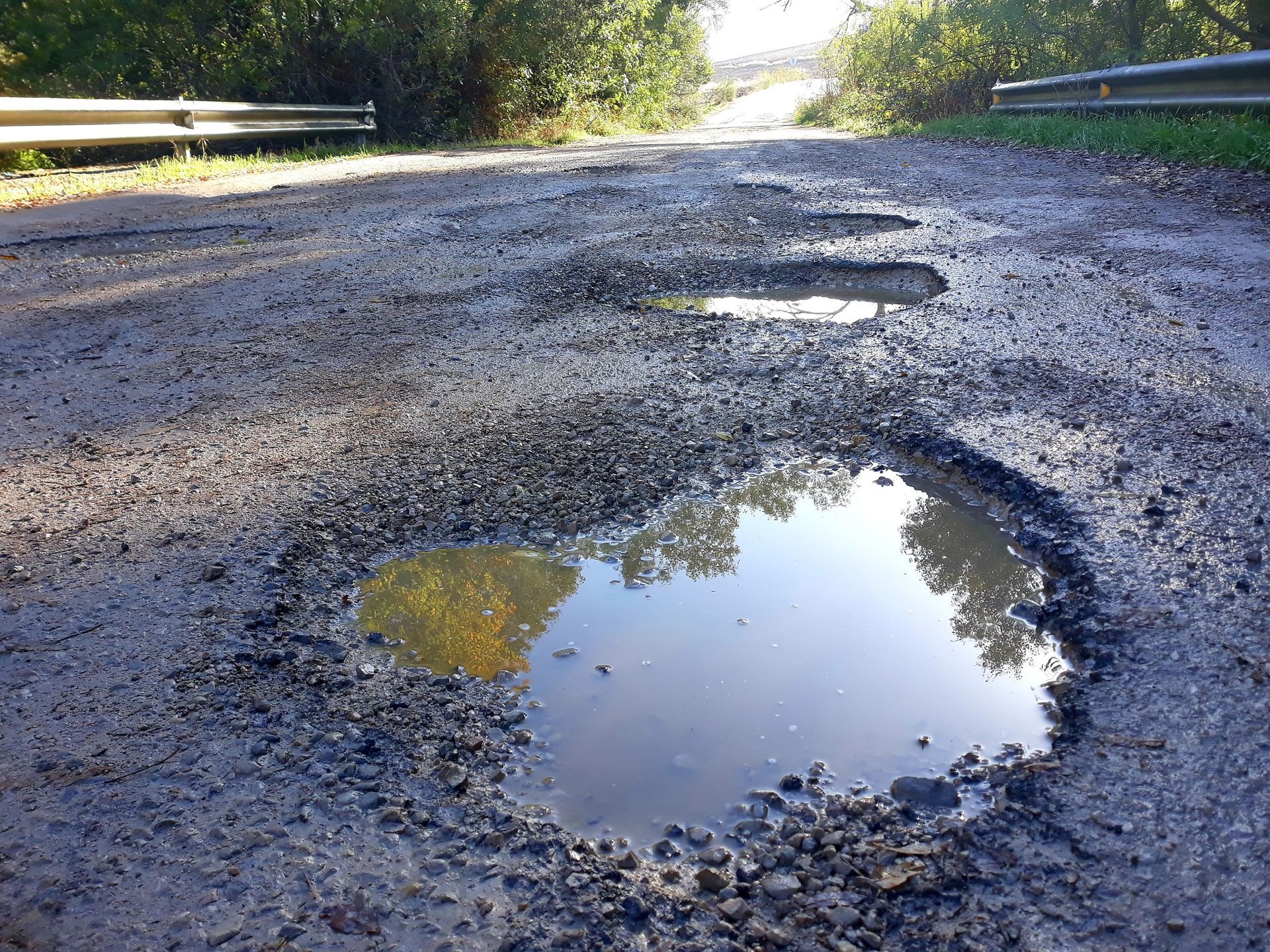 A gravel road with several large, deep potholes filled with rainwater, flanked by guardrails and trees.