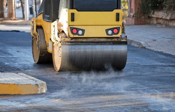 A yellow steamroller compacts fresh, steaming black asphalt on a road.