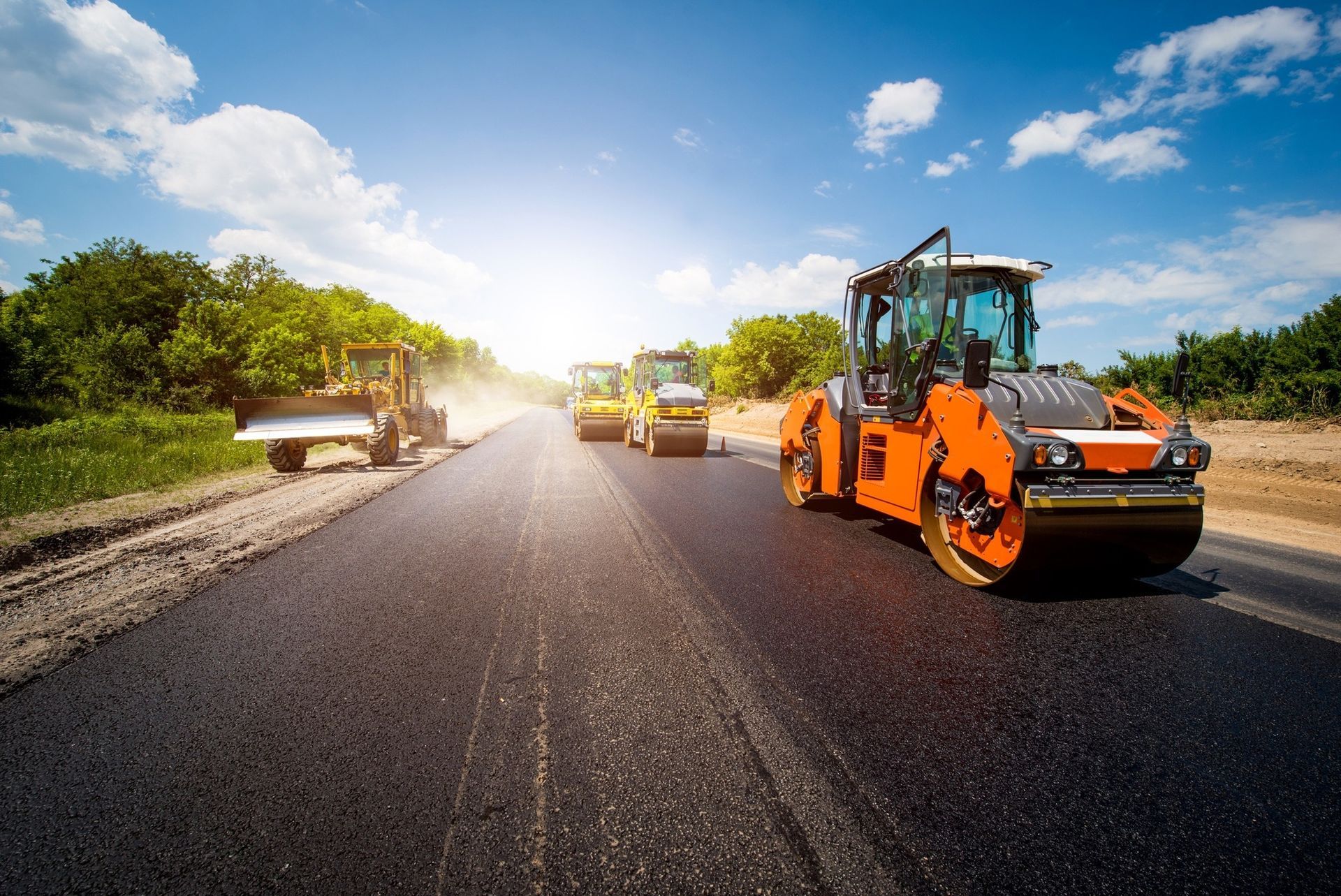 Bright orange rollers pave a new asphalt road under a sunny sky with green trees in the background.