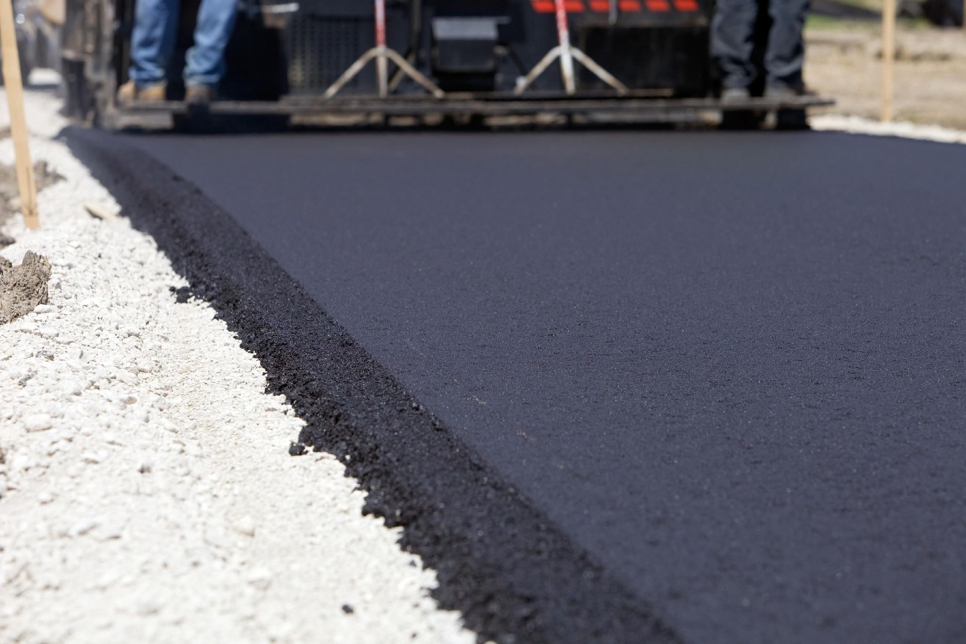 A construction paver lays fresh, dark asphalt on a road, flanked by light gravel, with workers visible in the background.