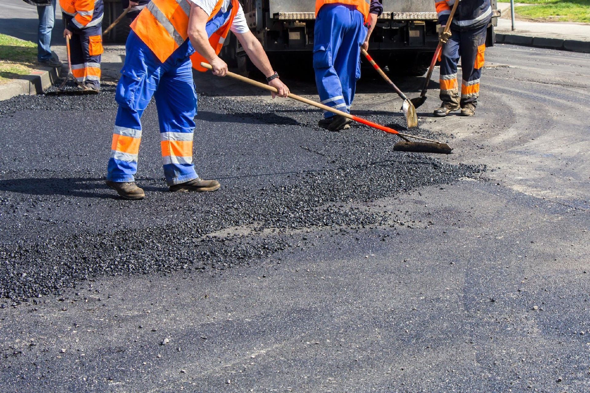 Construction workers in high-visibility orange vests and blue trousers use rakes to spread hot asphalt on a road.