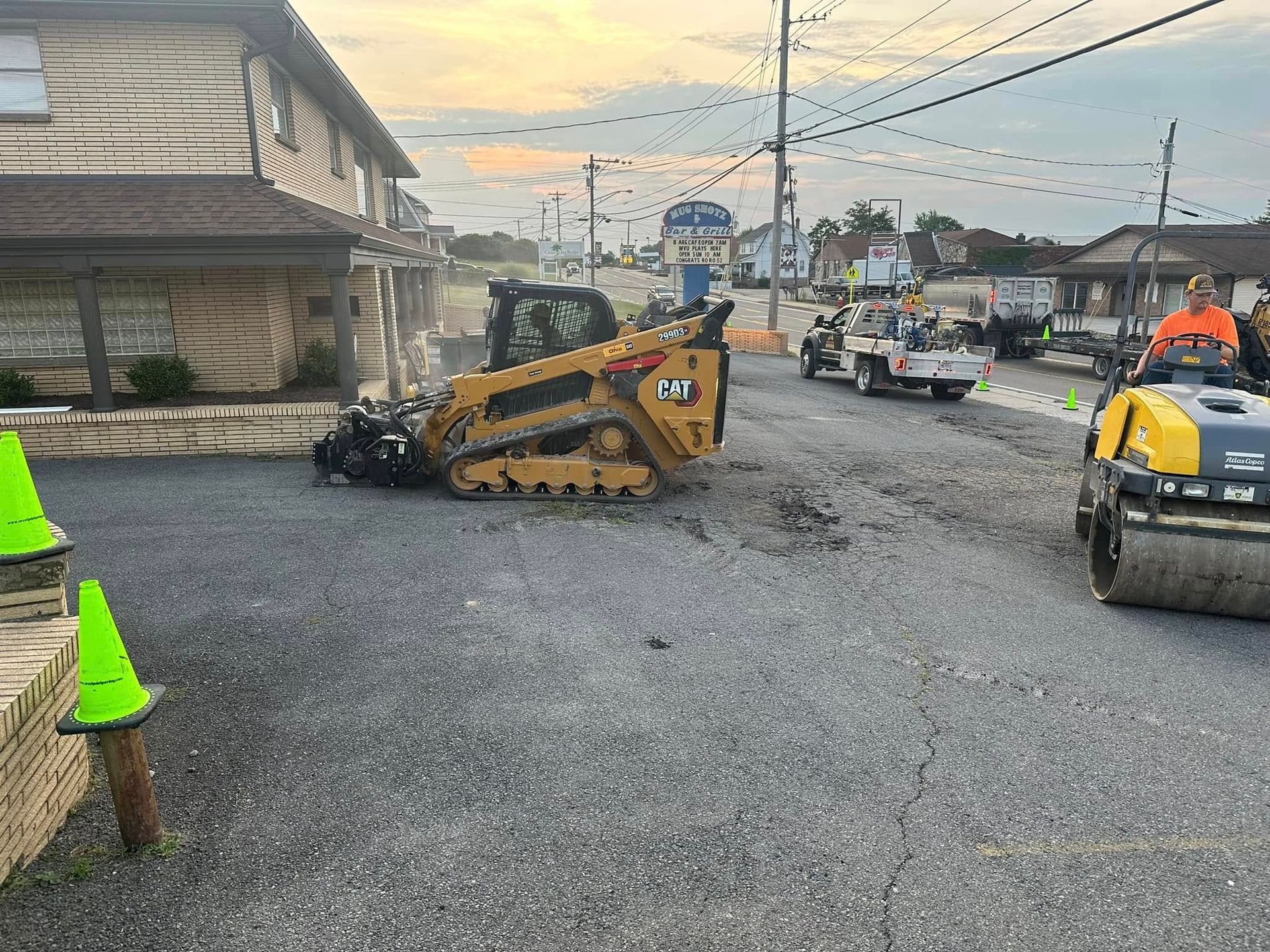 Construction equipment, including a yellow skid steer and a road roller, works on a gravel road next to a building.