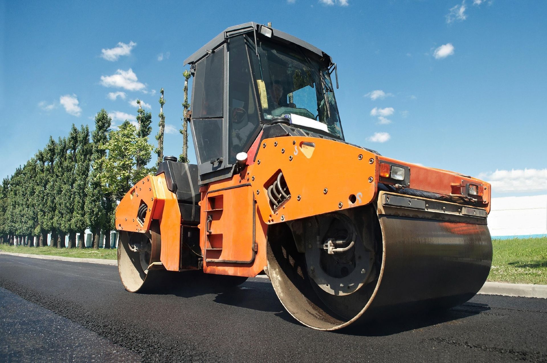 An orange road roller compacting freshly laid black asphalt on a sunny day next to a line of trees.