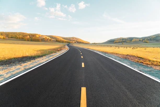 A paved road with a yellow center line stretches through a sunny, open landscape with rolling, golden hills.