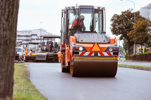 An orange road roller moves toward the camera on a paved road, with construction vehicles and workers in the background.