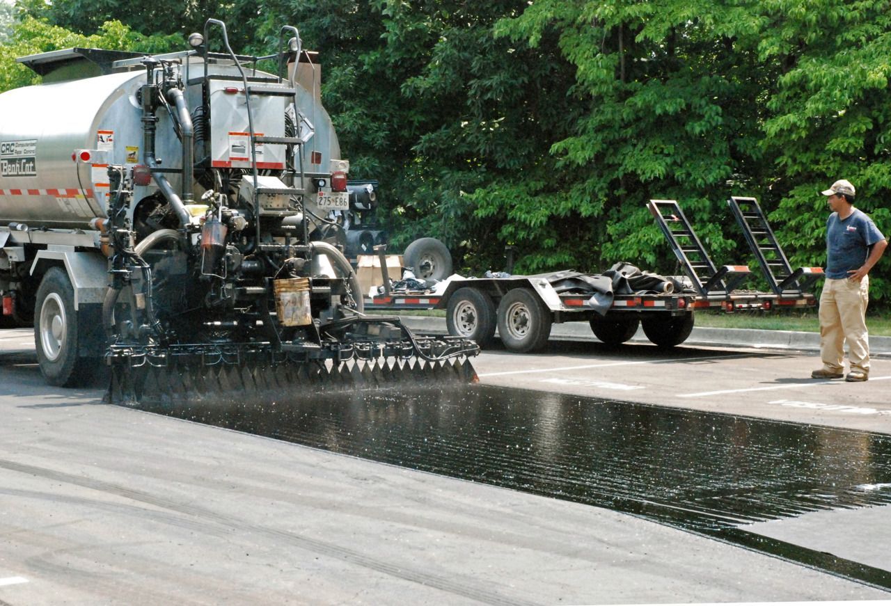 A truck sprays a layer of liquid asphalt onto a road surface as a worker stands nearby.