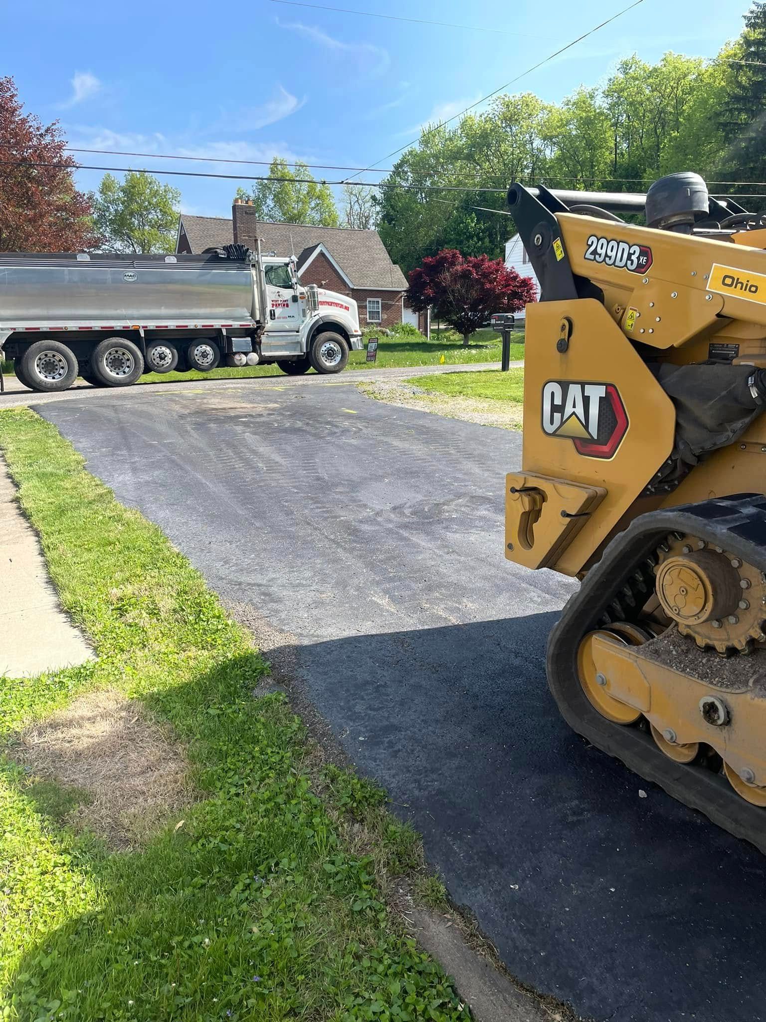 A yellow CAT skid-steer loader parked on a newly paved driveway with a dump truck nearby on a sunny day.