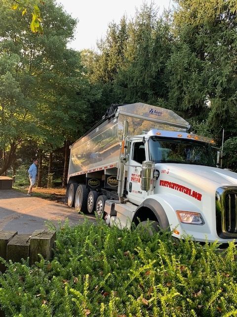 A white dump truck parked on a driveway near trees, with a person standing in the background.
