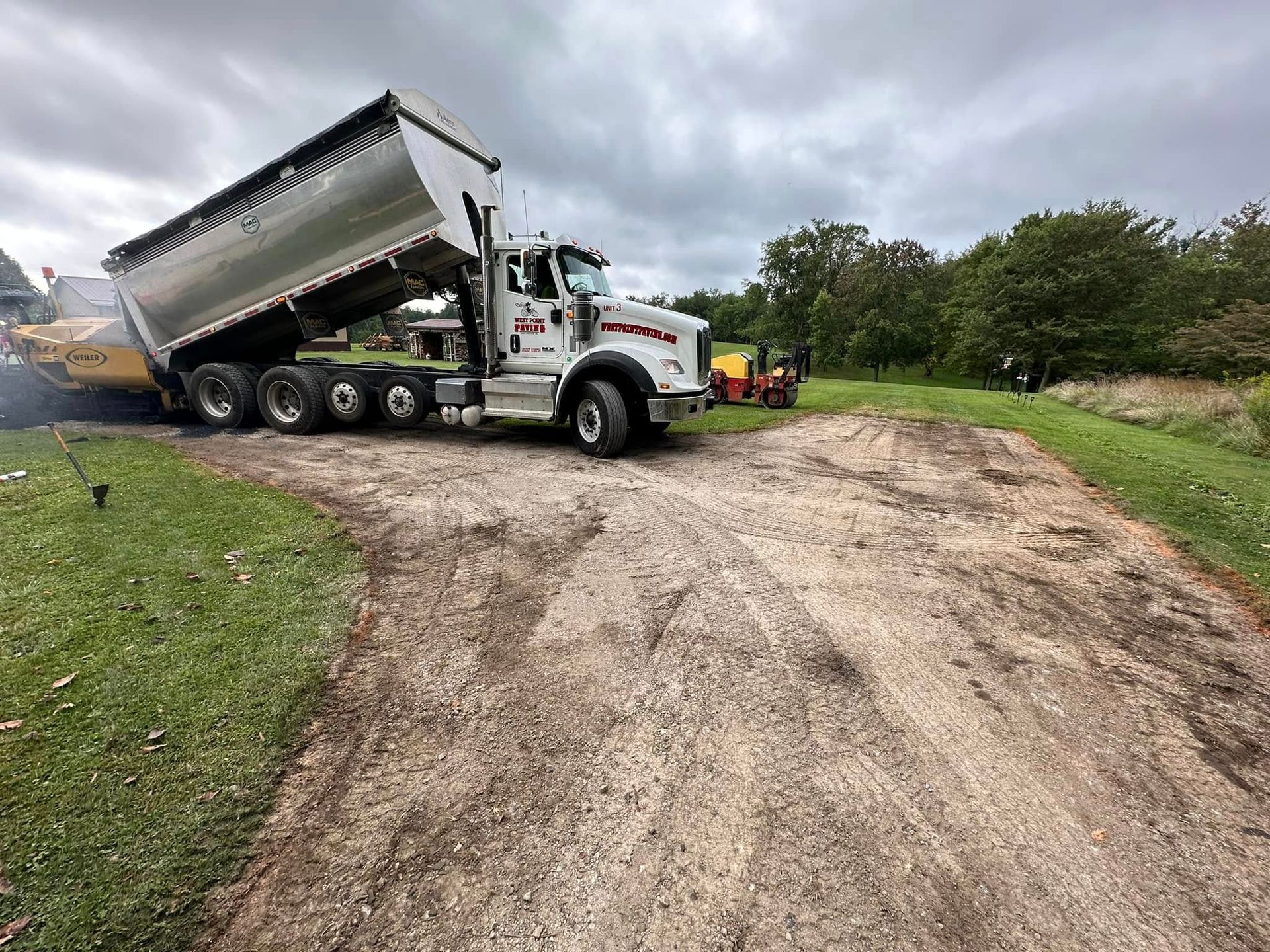 A white dump truck is raised and parked on a dirt path next to green grass and trees, with construction equipment nearby.
