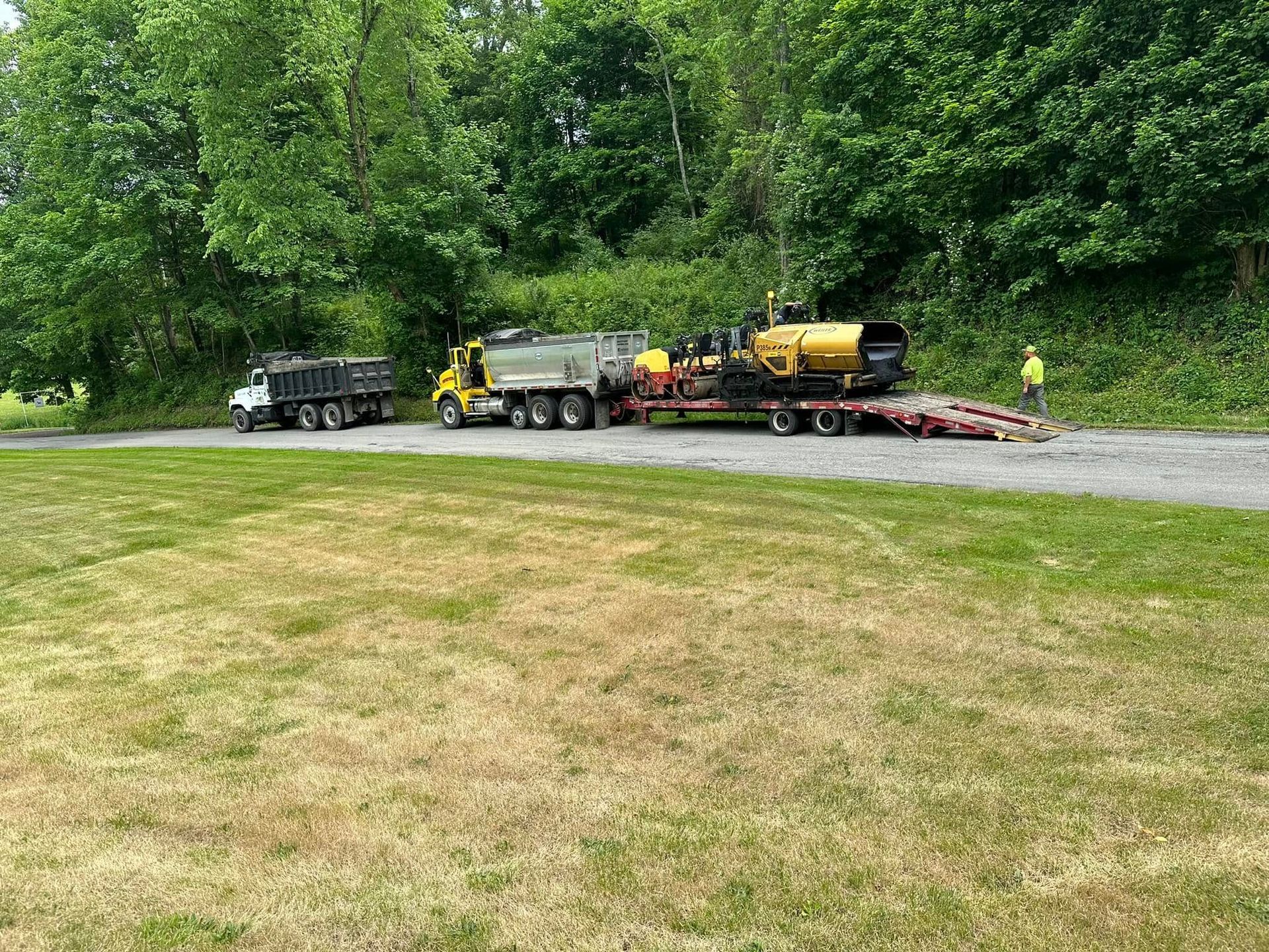 A yellow construction vehicle on a flatbed trailer pulled by a truck, with a dump truck nearby on a paved road.