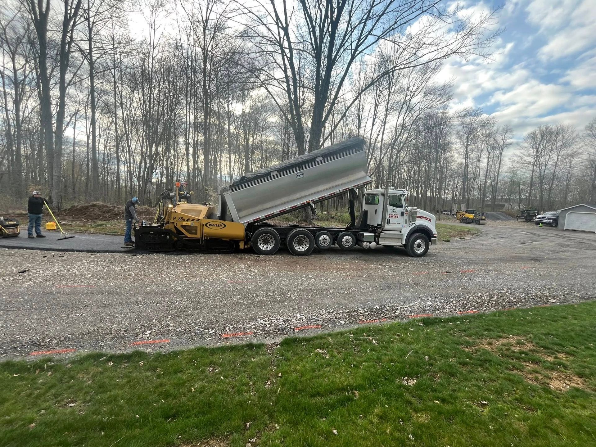 A dump truck pours asphalt into a road paver on a gravel lot in front of a tree line.