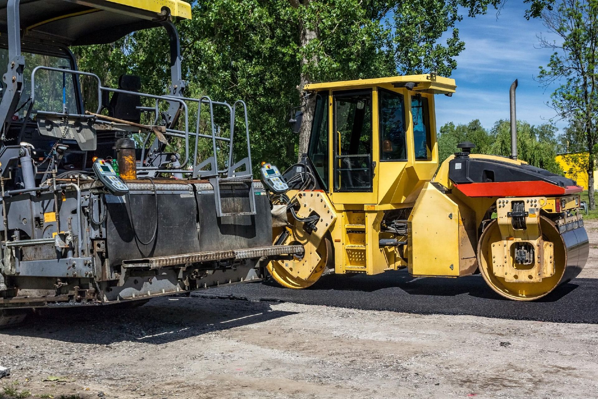 A yellow construction roller sits on freshly laid asphalt next to a large piece of heavy machinery in a wooded area.